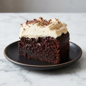 A rich slice of dark chocolate Guinness cake reveals a moist, deep-brown crumb. A generous, fluffy swirl of pale Irish buttercream tops the slice, garnished with delicate chocolate shavings. The cake sits on a simple dark plate, creating a dramatic contrast between the cake and frosting.