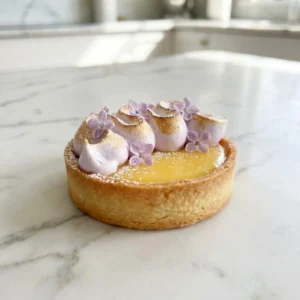 A delicate mini lemon tart with a golden, buttery crust and a glossy, vibrant yellow curd filling. The tart is crowned with beautifully torched, swirled meringue in a pale lilac color, garnished with fresh lilac blossoms and a fine dusting of powdered sugar.