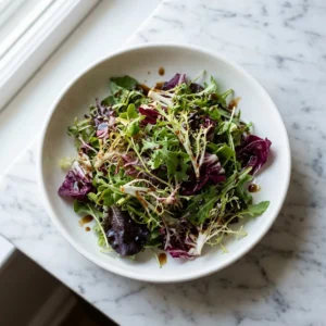 A vibrant spring mix salad featuring tender, multi-colored lettuces and deep purple radicchio. A glossy, dark balsamic honey dressing is generously drizzled over the top, making the fresh leaves glisten. The salad is served in a shallow, minimalist white ceramic bowl, showcasing its fresh colors.