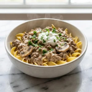 A rustic white bowl filled with creamy ground beef stroganoff over wide egg noodles. The rich, light brown sauce coats tender beef crumbles and mushroom slices. A dollop of white sour cream and a sprinkle of vibrant green parsley garnish the top, with gentle steam rising.