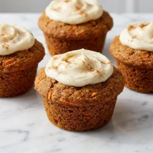 A close-up of rustic, golden-brown carrot cake muffins. The moist, dense crumb is studded with visible flecks of bright orange carrot. A thick, creamy white frosting is swirled on the domed tops, finished with a delicate dusting of cinnamon.