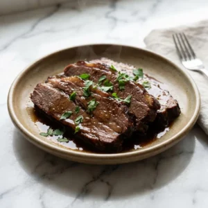 Thick, glistening slices of tender, fall-apart beef brisket rest in a pool of rich, dark jus on a rustic plate. The deep brown meat is garnished with vibrant fresh parsley, with a delicate wisp of steam rising from the dish.